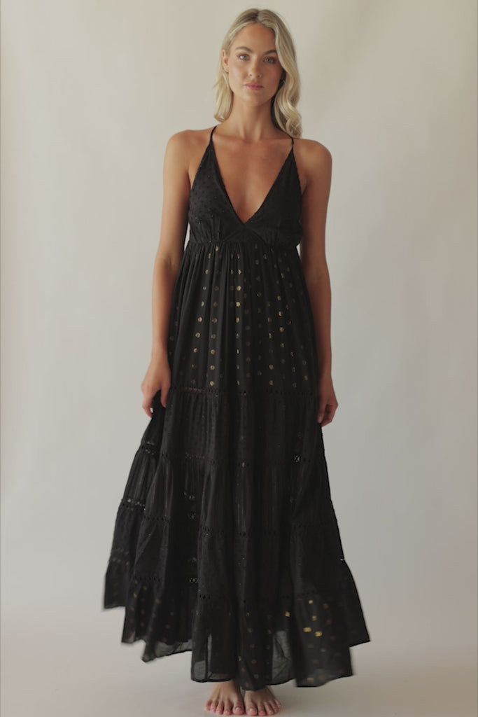 Blonde woman wearing a black full length dress with shimmery gold accents and cross stitching embroidery spinning around in front of a white wall.