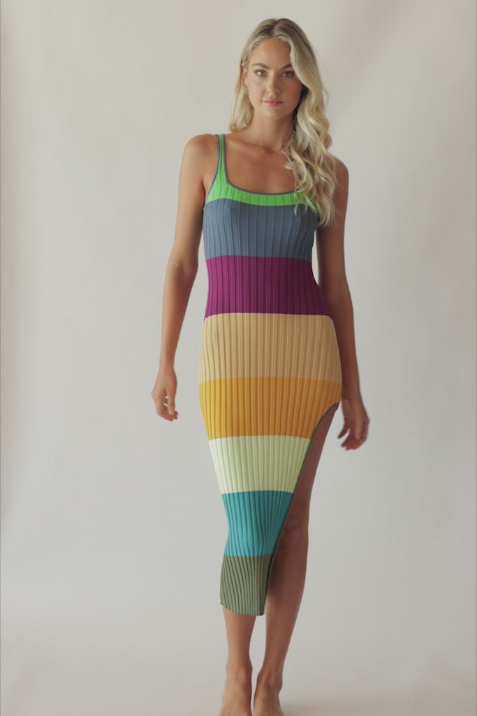 Blonde woman wearing a multi-colored stripe pattern dress with side slit spinning around in front of a white wall.