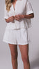 A woman wearing a white linen short sleeve shirt and shorts spinning against a white wall. 