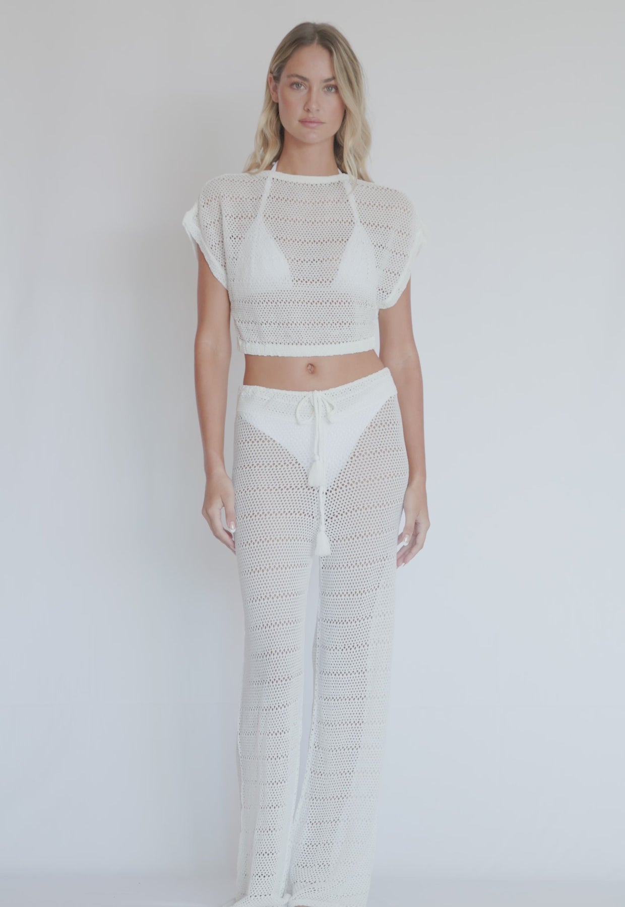 A blonde woman wearing a knit white crop top and long pants spinning in front of a white wall.