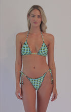 A blonde woman wearing a blue and green checkered bikini spinning in front of a white wall.