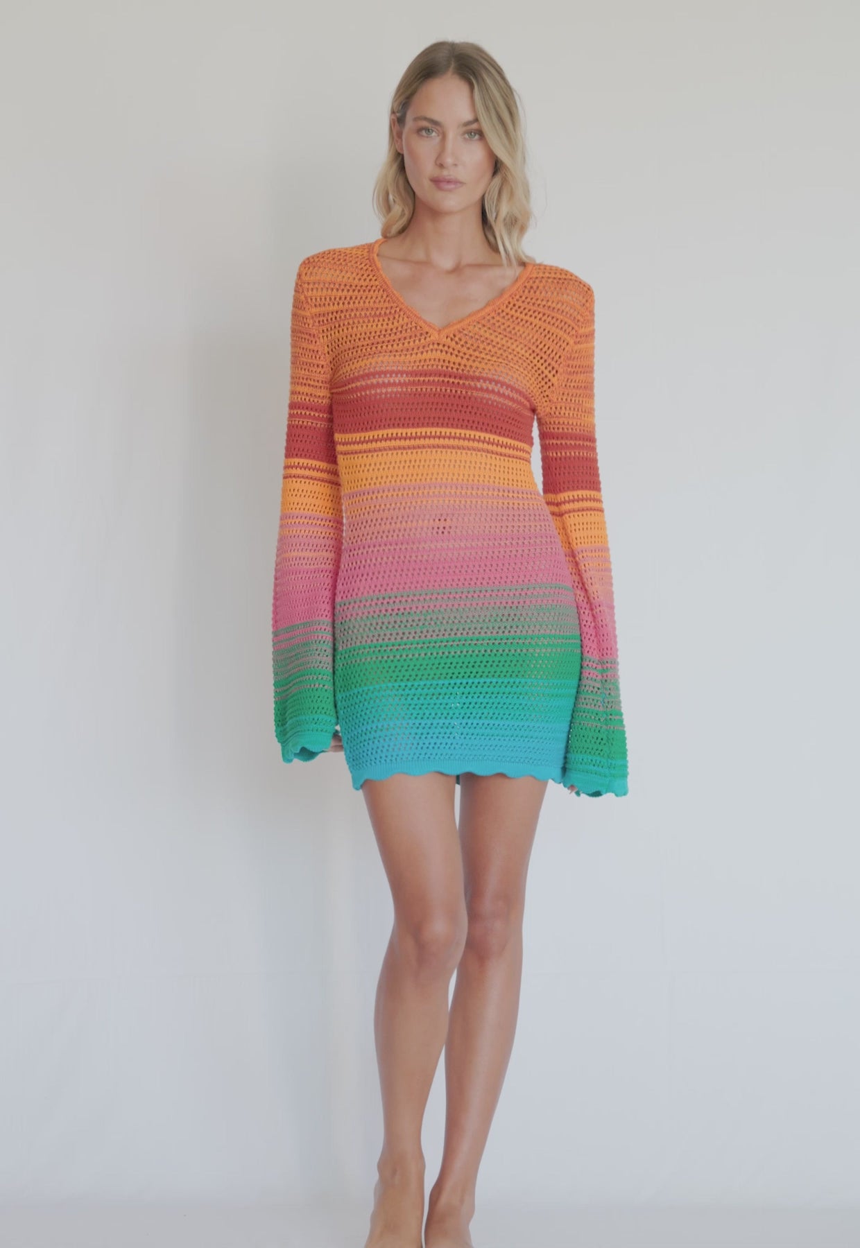 A woman wearing a long sleeve, knit, multi-colored dress spinning in front of a white wall.