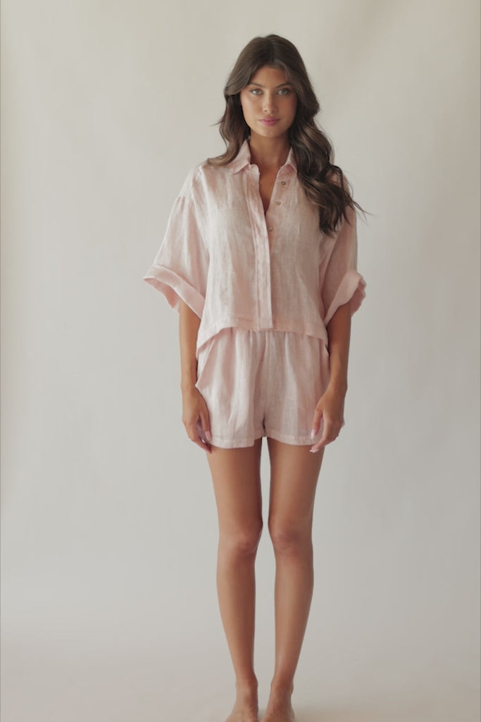 Brunette woman wearing a pink linen button coverup shirt and shorts spinning around in front of a white wall.