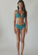 Brunette woman wearing a textured turquoise halter bikini with gold details spinning around in front of a white wall.