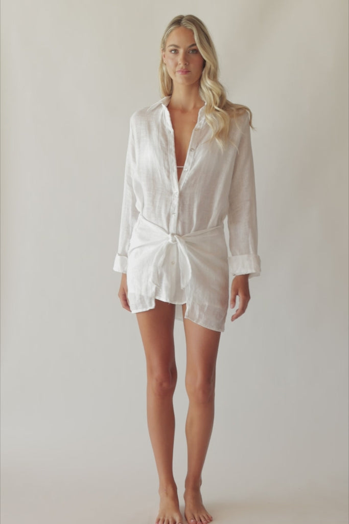 Blonde woman wearing a lightweight white linen button coverup with front-tie detail spinning around in front of a white wall.