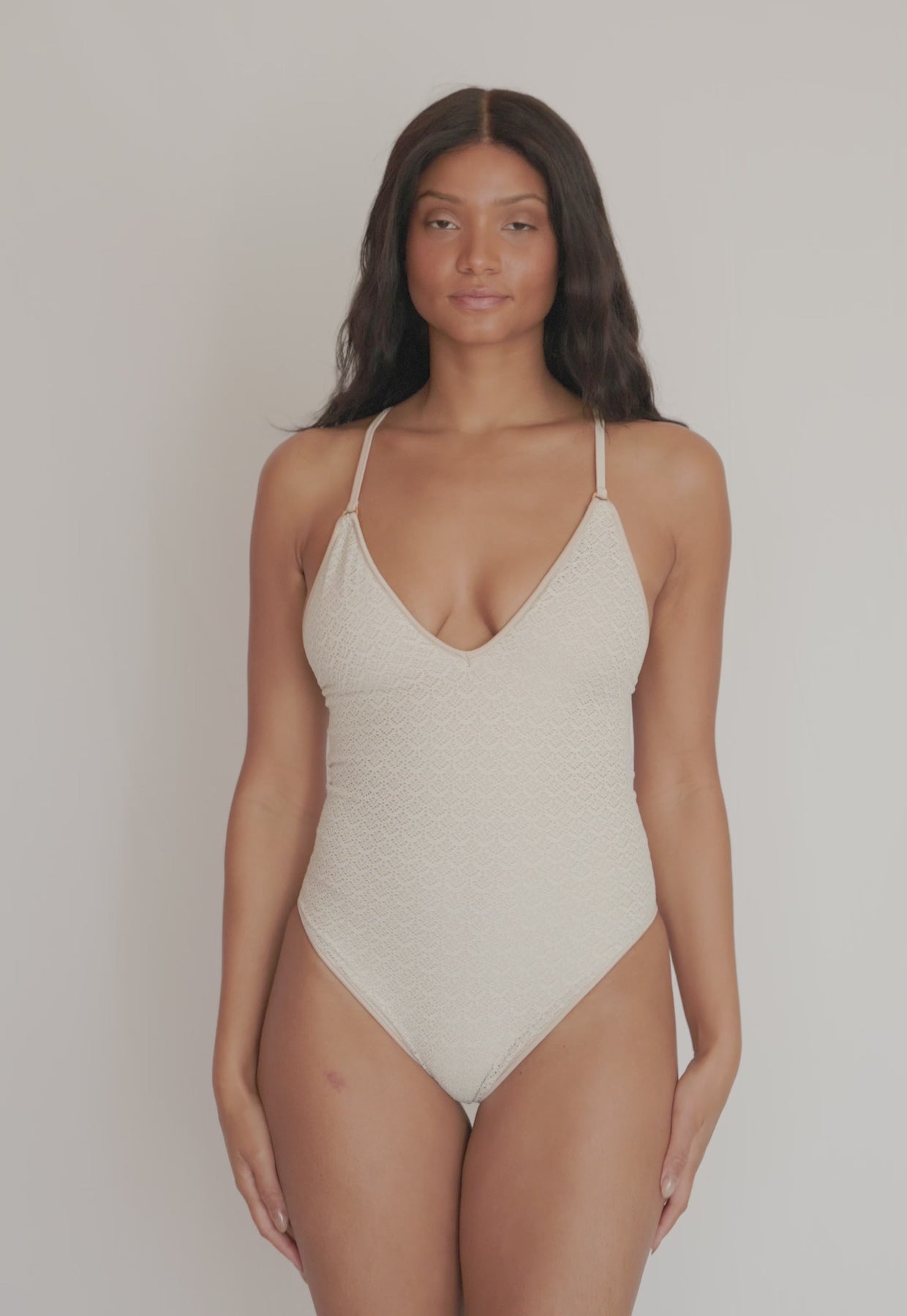 A woman wearing an Ivory one piece swimsuit spinning in front of a white wall. 