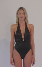 A blonde woman wearing a black one piece swimsuit spinning in front of a white wall. 
