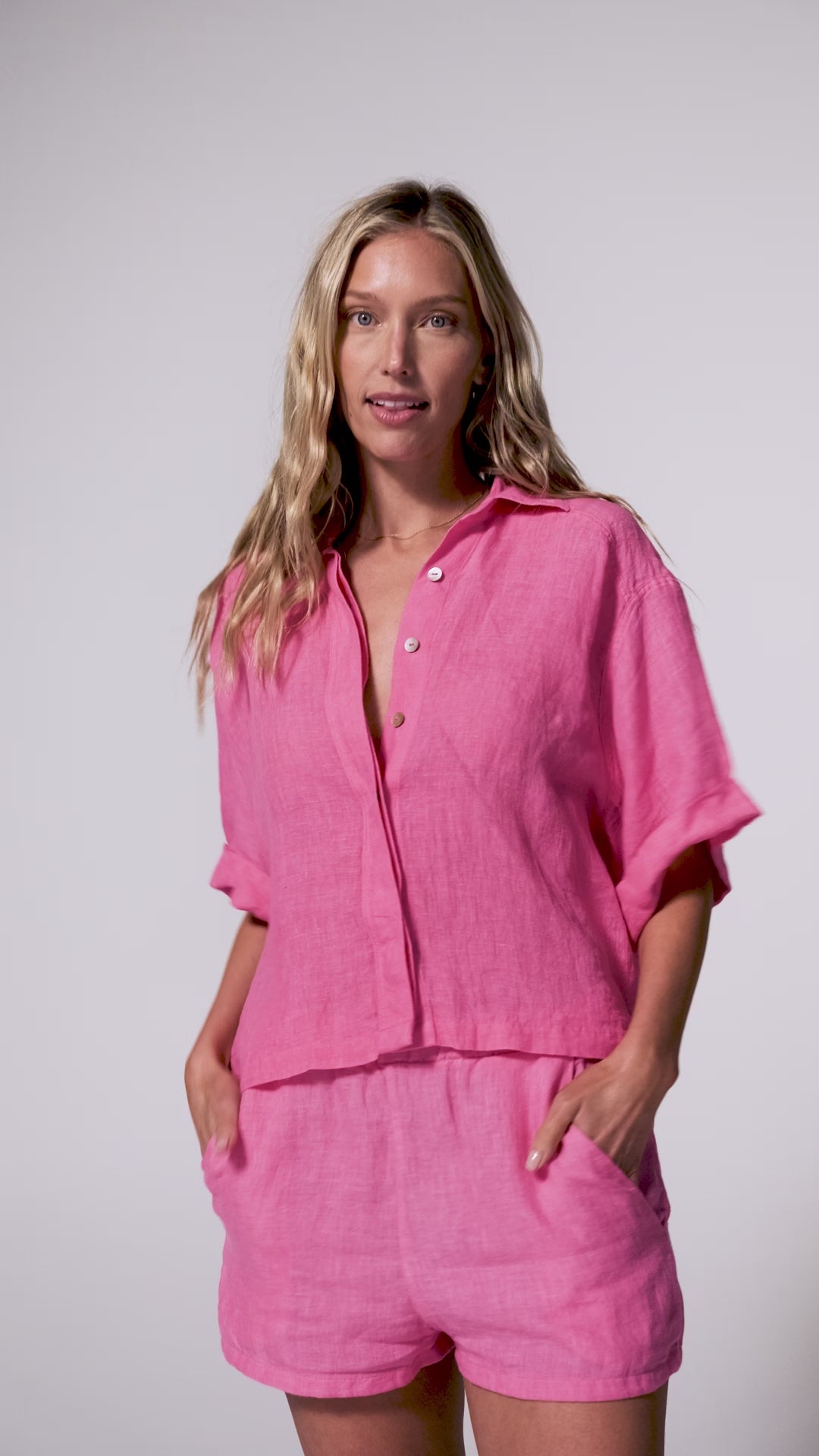 A woman wearing a bright pink linen shirt and matching shorts spinning in front of a white wall.