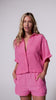 A woman wearing a bright pink linen shirt and matching shorts spinning in front of a white wall.