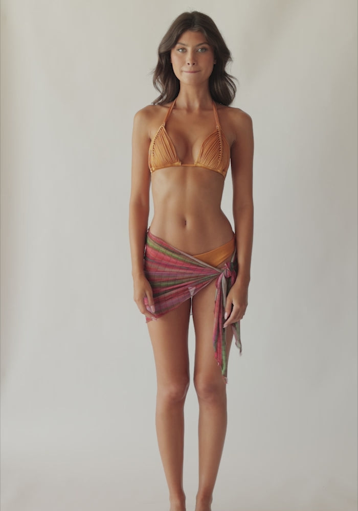 Brunette woman wearing an orange macrame triangle shape bikini and multi-colored stripe print sarong spinning around in front of a white wall.