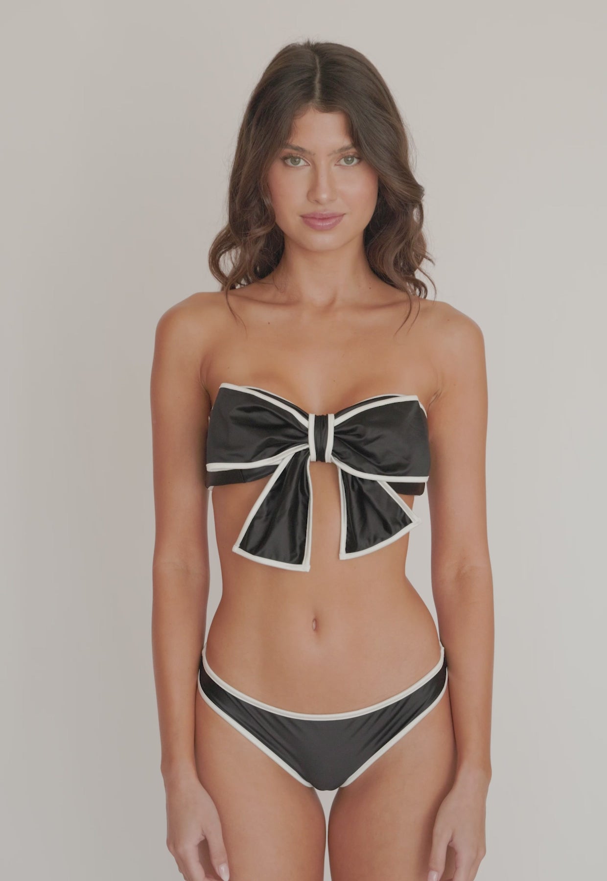 A woman wearing a black bandeau bikini with a bow detail spinning in front of a white wall. 