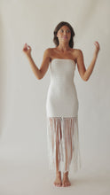 Brunette woman wearing a white strapless fringe hem dress with macramé details spinning around in front of a white wall.