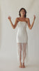 Brunette woman wearing a white strapless fringe hem dress with macramé details spinning around in front of a white wall.
