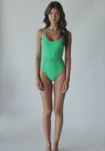 Brunette woman wearing green one piece swimsuit with gold beading spinning around in front of a white wall.