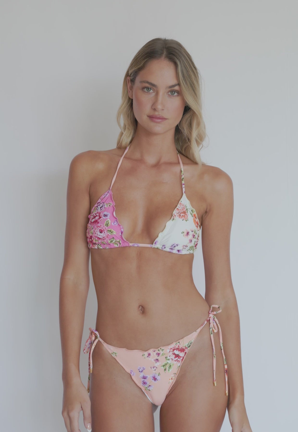 A woman wearing a floral triangle top bikini spinning in front of a white wall.