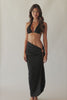 Brunette woman wearing a black halter bikini with net macramé details & shimmery gold accents and black skirt coverup with ring detail spinning around in front of a white wall.