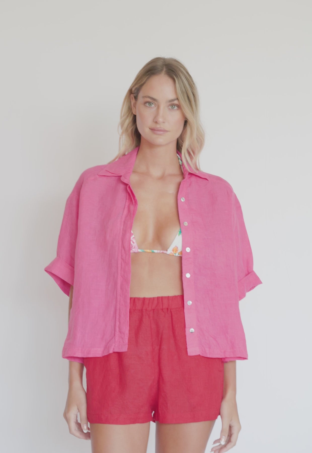 A blonde woman wearing a white floral bikini, red linen shorts, and a pink linen shirt spinning in front of a white wall. 