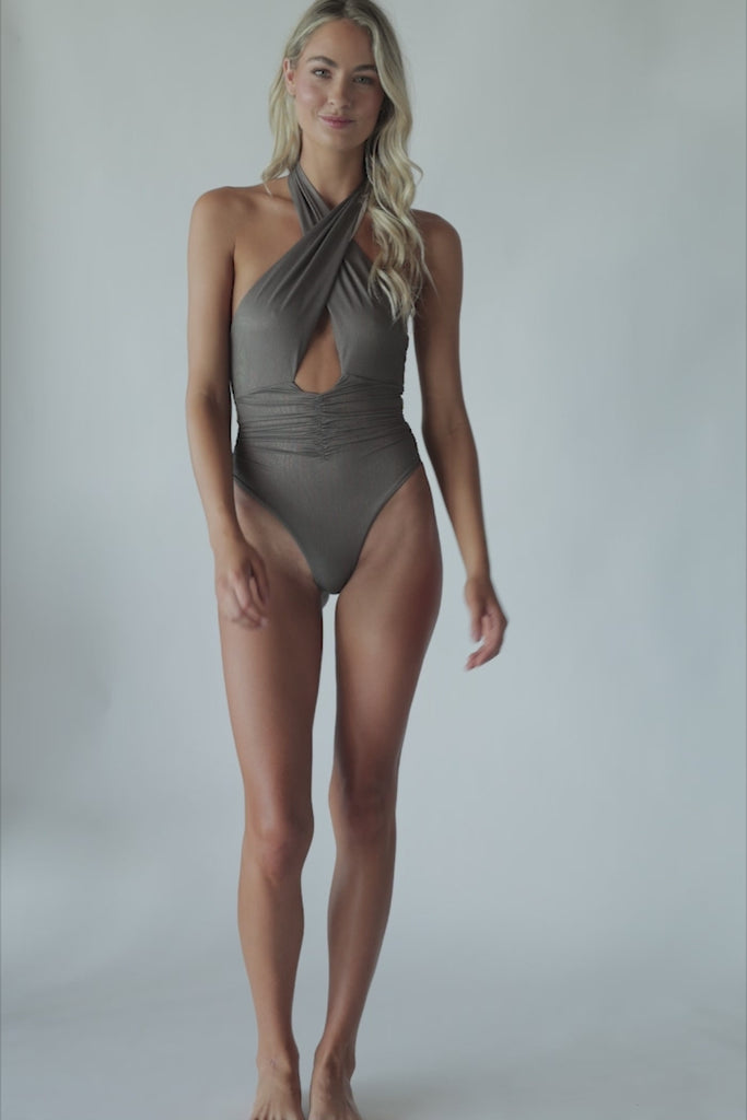 A blonde woman wearing a brown one piece bathing suit spinning in front of a white wall.