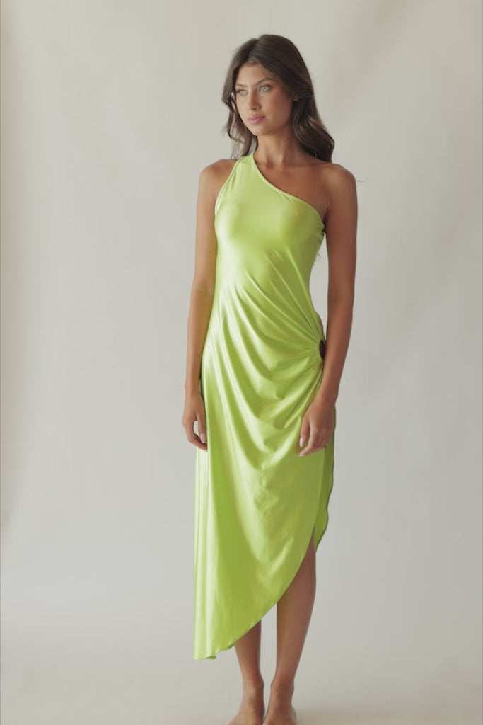 Brunette woman wearing a lime green one-shoulder dress with side slit and ring detail spinning around in front of a white wall.