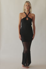 Blonde woman wearing a black full length coverup dress with stitching details and fringe spinning around in front of a white wall.