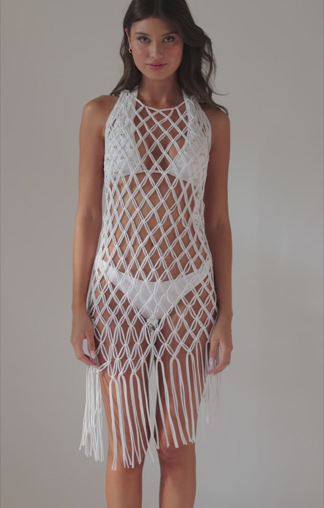 A brunette woman wearing a white bikini and white crochet dress spinning in front of a white wall.