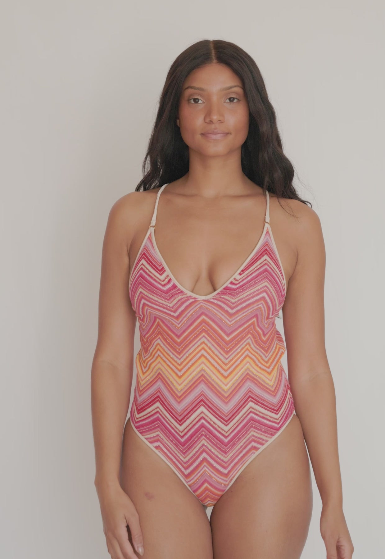 A woman wearing a pink and orange strip one piece swim suit spinning in front of a white wall. 