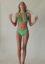 Blonde woman wearing a green halter bikini with net macramé & shimmery gold accents spinning around in front of a white wall.