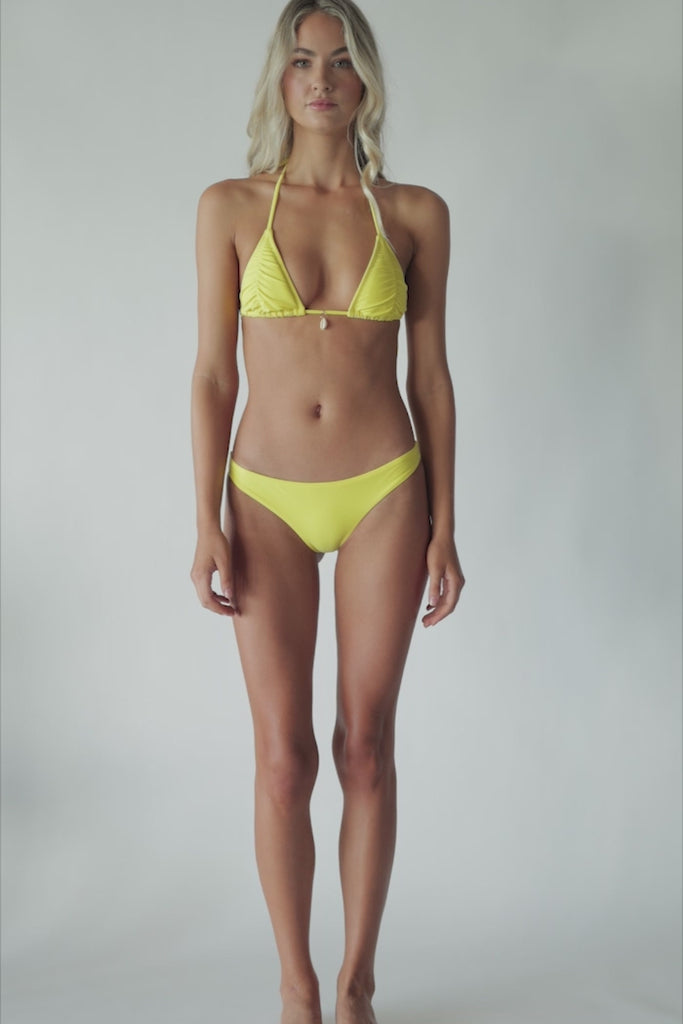 Blonde woman wearing a yellow ruched triangle top bikini spinning around in front of a white wall.