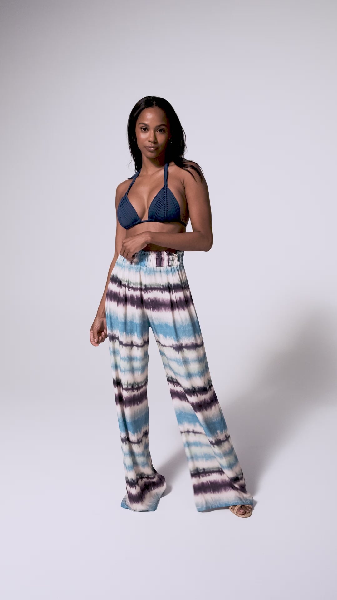 A woman wearing a navy triangle bikini top and blue tie dye pants spinning in front of a white wall.