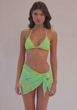 A brunette woman wearing a blue and green bikini and sarong spinning in front of a white wall. 
