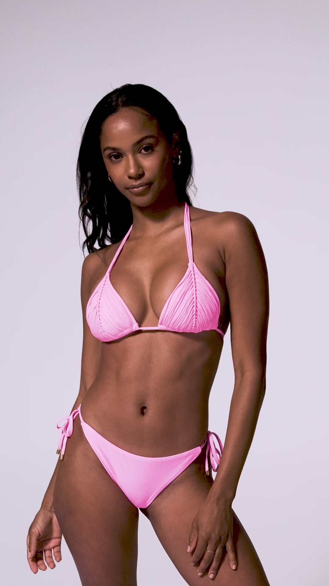 A woman wearing a pink triangle bikini top and matching tie side bottoms spinning in front of a white wall.