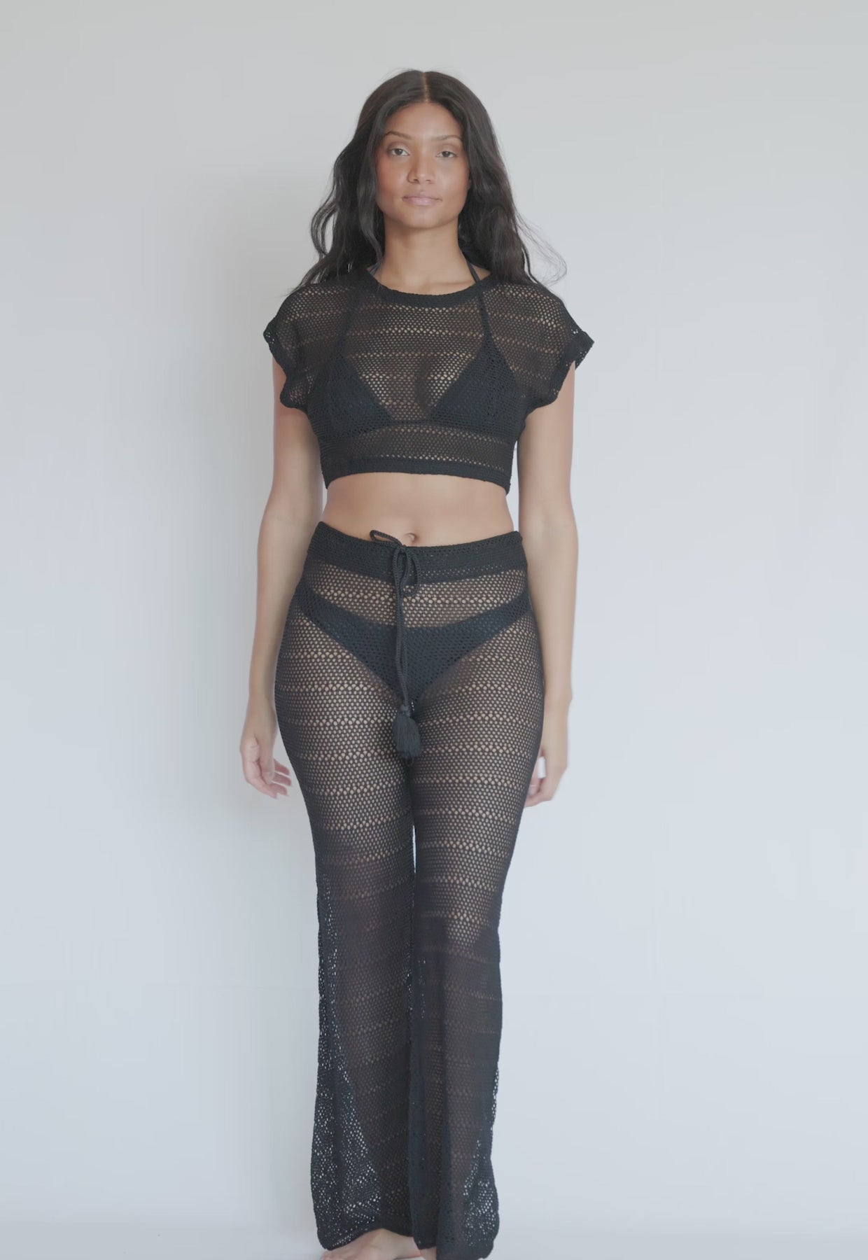 A woman wearing a black knit crop top and matching pants spinning in front of a white wall.