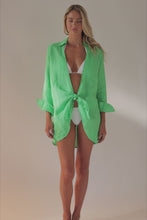 A blonde woman wearing a long sleeve green cover up over a white bikini spinning in front of a white wall.