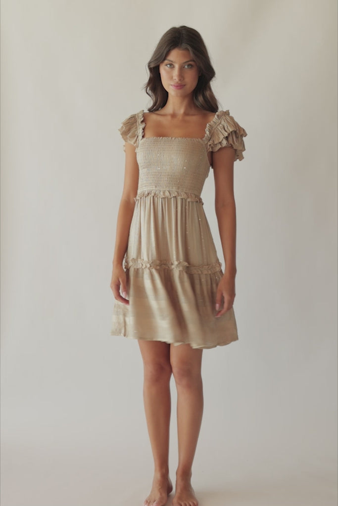 Brunette woman wearing a tan smocked top mini dress with ruffle straps and sequenced detailing spinning around in front of a white wall.