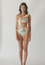 A brunette woman wearing a one piece with a tropical print spinning in front of a white wall. 