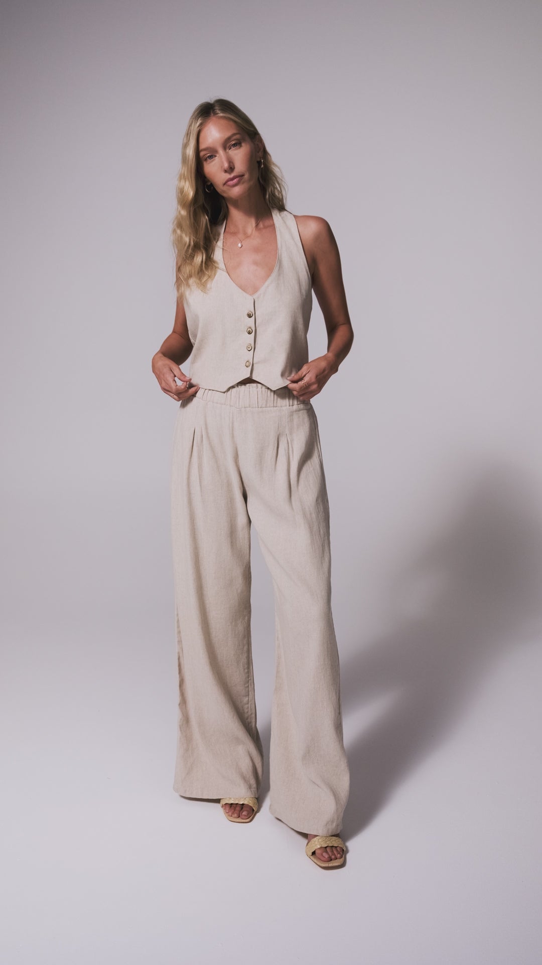 A woman wearing a tan vest and matching linen pants spinning in front of a white wall. 