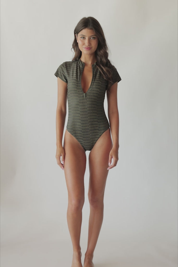 Brunette woman wearing a textured gray one piece swimsuit with zipper detail spinning around in front of a white wall.