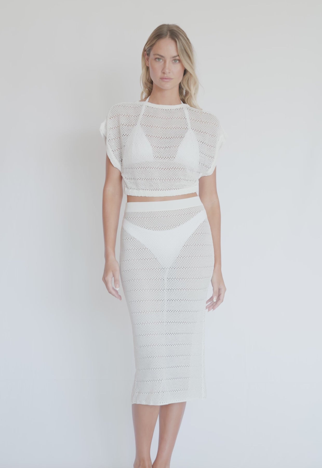 A blonde woman wearing a knit white crop top and midi skirt spinning in front of a white wall.