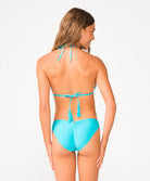 A woman wearing a turquoise triangle top bikini facing towards a white wall.