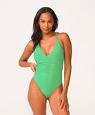 A woman wearing a green one piece swimsuit standing against a white wall. 